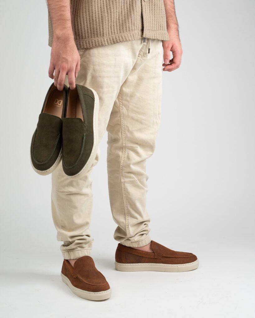 Hombre sosteniendo mocasines de gamuza verde mientras usa mocasines café combinados con pantalón claro en un estilo casual