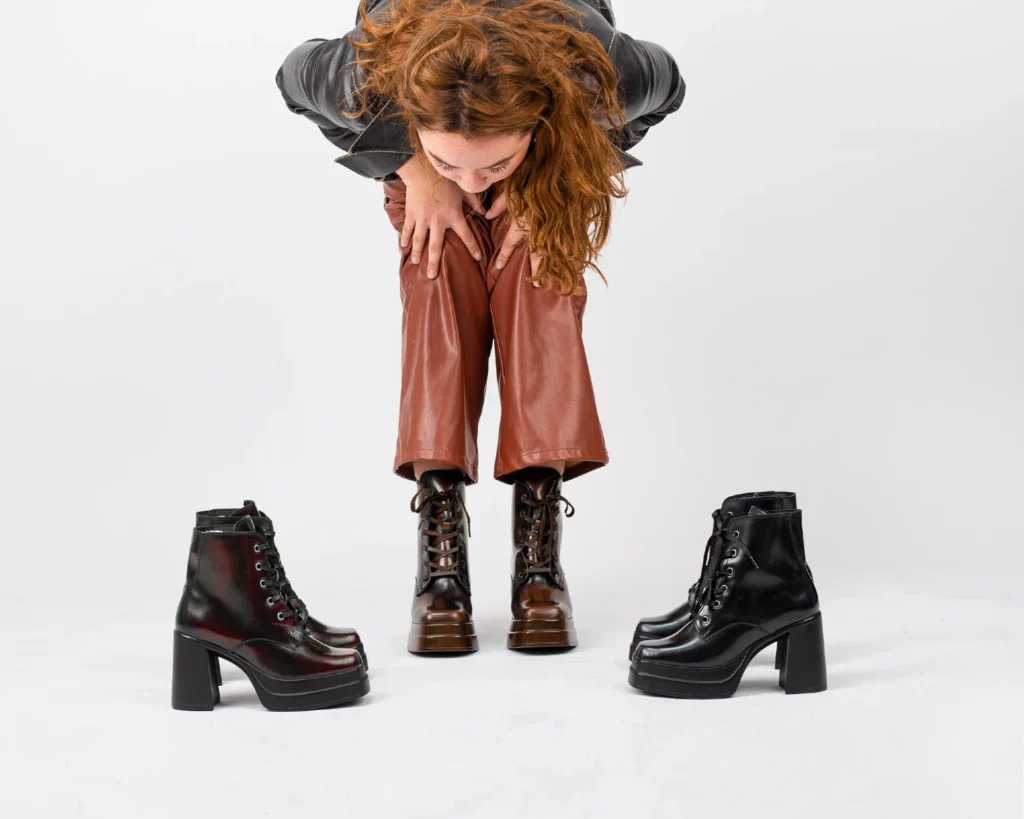 Mujer rodeada de botines negros, cafés y color vino como ejemplo de tres opciones complementarias para el guardarropa.