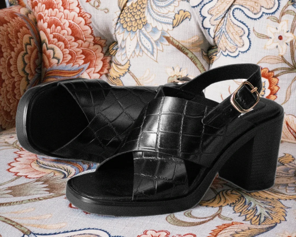 Primer plano de la sandalia de tiras cruzadas en negro con textura de cocodrilo, hebilla dorada y tacón bloque alto, reposando sobre un sillón con estampado floral. Muestra el diseño estructurado y versátil.