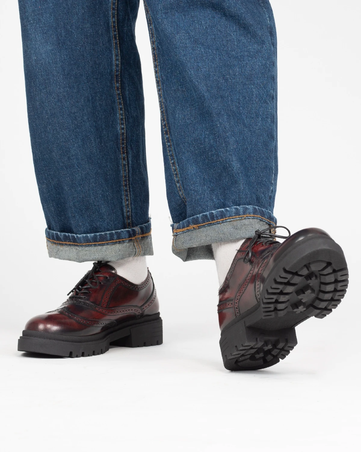Zapato moderno de vestir estilo bostoniano con picado brogue en tono vino y suela track robusta, combinado con jeans.