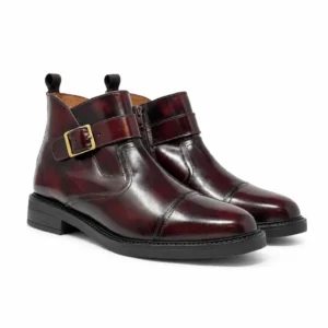 Par de botas elegantes para hombre elaboradas en piel color vino sombreado, con hebilla dorada y suela negra de perfil clásico.