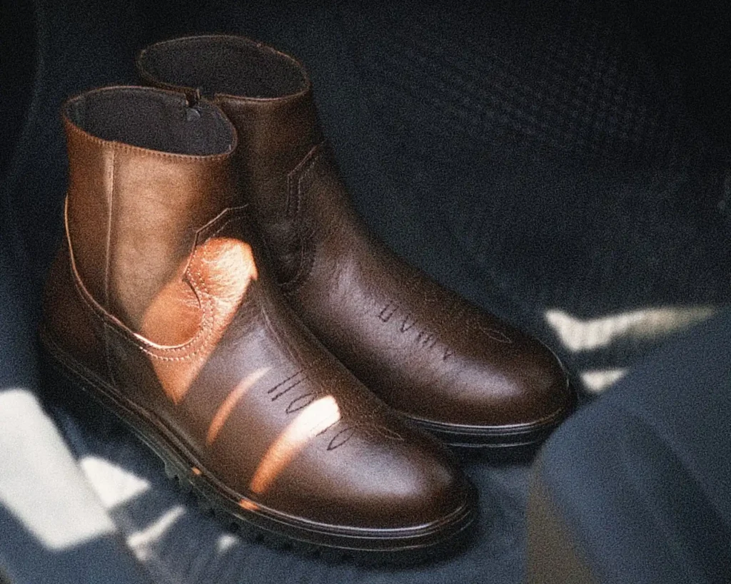 Par de botas Roper vaqueras para hombre en piel café, sin agujetas, con bordados en el empeine, cierre lateral y suela dentada negra.